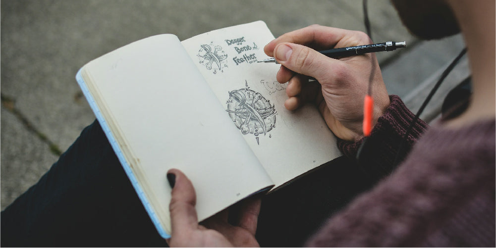 Photo by @koushikc. Image shows a person drawing with a pencil on a sketchbook.